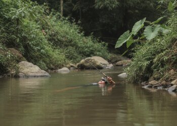 Gambar fotorealistik seorang pria yang ditemukan meninggal dunia, mengapung di tepi sungai pedesaan di Kulon Progo, dengan tangan masih memegang erat pancing.