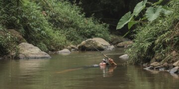 Gambar fotorealistik seorang pria yang ditemukan meninggal dunia, mengapung di tepi sungai pedesaan di Kulon Progo, dengan tangan masih memegang erat pancing.