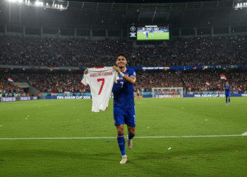Ole Romeny, a player from the Indonesian national football team, runs around the field of SUGBK, Jakarta, holding Marselino Ferdinan's jersey, in celebration of the team's victory in the FIFA ASEAN Cup 2026, while the crowd cheers and dramatic stadium lighting enhances the moment.