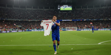 Ole Romeny, a player from the Indonesian national football team, runs around the field of SUGBK, Jakarta, holding Marselino Ferdinan's jersey, in celebration of the team's victory in the FIFA ASEAN Cup 2026, while the crowd cheers and dramatic stadium lighting enhances the moment.