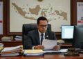 Seorang pria berpakaian seragam dan kacamata duduk di meja, di sekitarnya terdapat dokumen-dokumen politik dan komputer. Dia menatap dokumen di tangannya dengan ekspresi serius. Di latar belakang terl