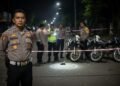 Sebuah foto jurnalistik profesional menampilkan seorang petugas kepolisian berdiri di depan lokasi kejadian di Makassar. Latar malam dengan cahaya lampu jalan yang redup menciptakan suasana yang seriu