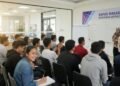 Sebuah kelompok siswa usia remaja, mungkin tingkat SMA atau perguruan tinggi, duduk dengan fokus di kelas atau ruang seminar. Ruangan terlihat terang, dengan papan tulis putih besar di depan yang mena
