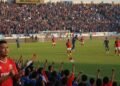 Oknum Dishub Makassar Diamankan di Polres Gowa A packed soccer stadium during a match between PSS Sleman and PSM Makassar. The atmosphere is lively with spectators in blue and red, the colors of the teams. The game is intense with players in actio