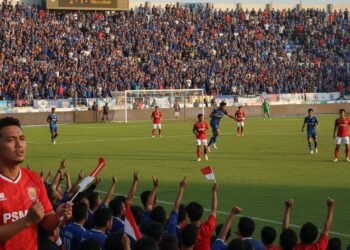 A packed soccer stadium during a match between PSS Sleman and PSM Makassar. The atmosphere is lively with spectators in blue and red, the colors of the teams. The game is intense with players in actio