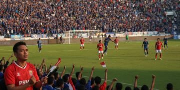 A packed soccer stadium during a match between PSS Sleman and PSM Makassar. The atmosphere is lively with spectators in blue and red, the colors of the teams. The game is intense with players in actio