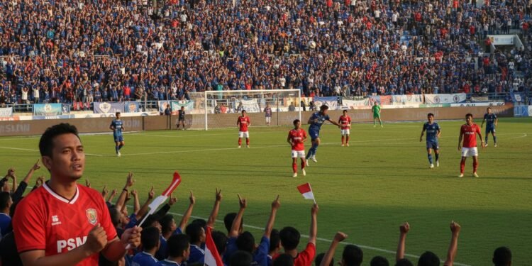 Prediksi Skor PSS Sleman Vs PSM Makassar: Peluang Menang Juku Eja 20 Persen A packed soccer stadium during a match between PSS Sleman and PSM Makassar. The atmosphere is lively with spectators in blue and red, the colors of the teams. The game is intense with players in actio