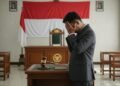 Seorang pria muda berpakaian seragam, kemungkinan MA. Untung, berdiri di depan meja hakim dengan ekspresi cemas dan memikirkan, menahan kepalanya dengan tangan. Ruangan pengadilan yang terang dengan b