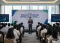 H. Gunadi, the keynote speaker at IRIS 2027, addresses a group of young people in a modern conference hall. The hall is filled with a diverse audience, all engaged in the event. A large banner with th