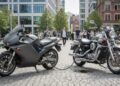 Sebuah motor listrik modern dengan port pengisian daya terlihat di depan, dibandingkan dengan motor bensin tradisional yang menunjukkan mesin dan emisi. Latar belakang menampilkan kota dengan beberapa