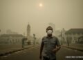 Makassar Targetkan 9.885 Kasus TBC Sembuh dalam Tiga Tahun, Apa Strateginya? Budi S., seorang warga Makassar yang memakai masker, berjalan di jalanan yang terpapar asap, di depan Alun-Alun Makassar yang tampak kabut. Pada latar belakang, matahari tampak pudar dan berwarna kuni