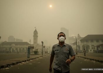 Budi S., seorang warga Makassar yang memakai masker, berjalan di jalanan yang terpapar asap, di depan Alun-Alun Makassar yang tampak kabut. Pada latar belakang, matahari tampak pudar dan berwarna kuni
