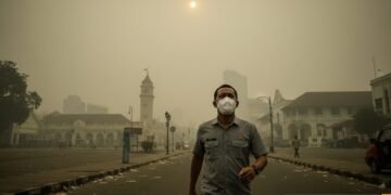 Budi S., seorang warga Makassar yang memakai masker, berjalan di jalanan yang terpapar asap, di depan Alun-Alun Makassar yang tampak kabut. Pada latar belakang, matahari tampak pudar dan berwarna kuni