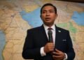 Budi S., Walikota Makassar, berdiri dengan percaya diri di sebuah ruangan modern dengan latar belakang layar besar UN di belakangnya. Dia berbicara di depan mikrofon dengan tangan yang bergerak-gerak,