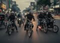 Sebuah jalan di Makassar yang ramai pada sore hari, dengan orang-orang berjalan dan kendaraan berlalu. Tiba-tiba, sekelompok pengendara motor, berpakaian hitam dan memakai helm, muncul dari keramaian.
