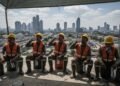 Sekelompok pekerja konstruksi di Jakarta beristirahat di bawah area berbayang pada hari yang terik. Mereka berkeringat deras dan tampak lelah, masih memakai topi aman dan jaket kerja. Matahari bersina