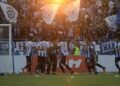 PSIM Juku Eja players celebrate their 2-1 victory against PSM Makassar with excited fans in the stadium. The setting sun gives a warm glow to the scene, and the crowd is cheering and waving flags.