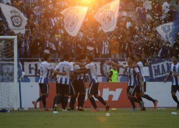 PSIM Juku Eja players celebrate their 2-1 victory against PSM Makassar with excited fans in the stadium. The setting sun gives a warm glow to the scene, and the crowd is cheering and waving flags.