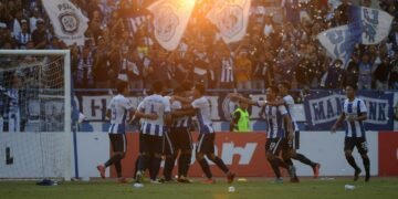 PSIM Juku Eja players celebrate their 2-1 victory against PSM Makassar with excited fans in the stadium. The setting sun gives a warm glow to the scene, and the crowd is cheering and waving flags.
