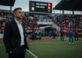 Van Gastel, yang tampak berpikir, berdiri di sisi lapangan stadion, memperhatikan hasil pertandingan. Latar belakang menunjukkan papan skor dengan skor PSIM di posisi lebih rendah dibandingkan PSM Mak