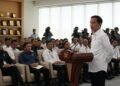 Presiden Joko Widodo berdiri di podium di ruang konferensi yang luas dan modern, dengan tangan di belakang punggungnya dan menatap audiens dengan ekspresi yang tegas dan tenang. Ruangan terang dengan