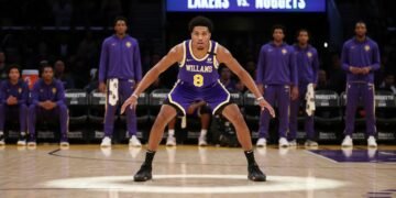 Ziaire Williams, a confident basketball player, stands on the court with the Lakers' bench area in the background, ready to start the game against the Nuggets. The stadium lights create a focused and