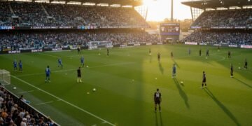 Stadion sepak bola yang penuh dengan penonton yang antusias, pemain Melbourne Victory dalam kostum rumput mereka berlatih di lapangan, sementara pemain Wellington Phoenix memakai kostum tandang. Penon