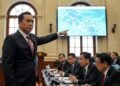 Pilwali Makassar Terancam Ditunda MA. Untung, an MP, is standing in a parliamentary chamber, gesturing towards a map on a screen, expressing concern about the proposed Kopdes for salary distribution. The background shows other legisla