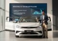 M. Ridham standing in front of a BYD Atto 2 electric vehicle in a modern dealership. The car is shown with its LED headlights and dashboard visible. A large digital screen in the background displays r