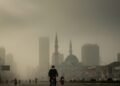 Dinas Perpustakaan Makassar Perkenalkan Minat Baca Sistem Inklusi Sosial A dense fog covers the city of Makassar-Sinjai, with buildings barely visible. A cyclist wearing a mask rides along a busy street, with the city's iconic mosque minarets in the background. The sky is