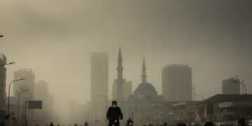 A dense fog covers the city of Makassar-Sinjai, with buildings barely visible. A cyclist wearing a mask rides along a busy street, with the city's iconic mosque minarets in the background. The sky is