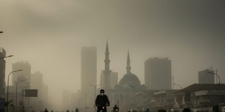 Kualitas Udara Makassar-Sinjai 18 Oktober: Status Tidak Sehat Akibat PM2.5 A dense fog covers the city of Makassar-Sinjai, with buildings barely visible. A cyclist wearing a mask rides along a busy street, with the city's iconic mosque minarets in the background. The sky is