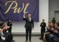 Andi Sudirman menerima penghargaan dari PWI Sulsel. Dia berdiri dengan percaya diri di atas panggung dengan trofi di tangan. Penonton duduk dalam barisan dengan tatapan penuh penghargaan. Penerangan y