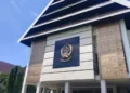 Gubernur Budi S. menghadiri parade sehat dan kirab swasembada pangan di Makassar, diiringi warga yang bersemangat dan bendera berwarna-warni. Latar belakang menampilkan campuran rumah tradisional dan