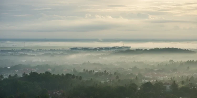 Denpasar, Bali, berawan, dengan Ngurah Rai International Airport sebagai objek utama. Latar belakang ditutupi oleh kabut tipis, menciptakan suasana tenang. Langit berwarna abu-abu lembut dengan awan p