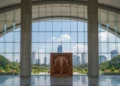 A grand entrance of the DPR building in Jakarta, with a sleek glass facade and ornate door, reflecting sunlight. The interior is bright with large windows, and the background shows greenery of the sur