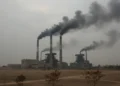 Kopdes Merah Putih “Cicil” Sebelum Beroperasi, Menkop Ferry Juliantono Ditekan DPR A depiction of an industrial complex with multiple smokestacks emitting dark smoke, symbolizing high levels of air pollution in Muaro Jambi. The sky is overcast, and the ground is covered with dust, w
