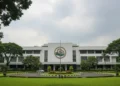 BMKG's main building in Jakarta is shown with its modern, white exterior and prominent logo. The building is surrounded by greenery, and the sky is overcast with light clouds, suggesting potential rai