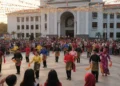 Sebuah taman kota yang ramai dengan orang-orang menonton dan berpartisipasi dalam penari joget yang menari. Bangunan pemerintah modern dengan detail arsitektur yang indah menjadi latar belakang, menan