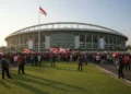 Gedung Stadion Nasional Jakarta dengan arsitektur modern dan nafas dinamis, dihiasi dengan bendera nasional yang berpendar. Lingkungan sekitarnya dipenuhi oleh keramaian penggemar sepak bola dan warta