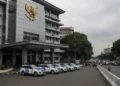 Lanskap luar Gedung Polda Sulsel yang modern dengan fasad kaca dan emblema polisi di depan pintu masuk utama. Lingkungan sekitarnya penuh dengan jalan raya yang sibuk, pejalan kaki, dan kendaraan, men