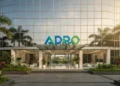 The main building of PT Adaro Energy Tbk is shown with a sleek glass facade and a prominent ADRO logo. The surroundings are green and well-kept, with bright natural light illuminating the entrance. Th