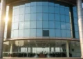PJ Sekda Makassar Hadiri Sertijab Komandan Lantamal VI Makassar The main building of BPKAD in Makassar is shown with a modern glass facade reflecting sunlight. The entrance is flanked by imposing pillars, and the interior is spacious with large windows. The scene