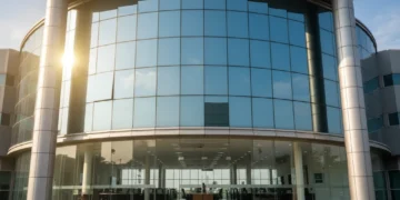 The main building of BPKAD in Makassar is shown with a modern glass facade reflecting sunlight. The entrance is flanked by imposing pillars, and the interior is spacious with large windows. The scene