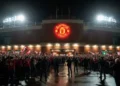 The Old Trafford stadium at night, with its red-brick facade and illuminated entrance, surrounded by excited fans and media. The Manchester United crest is prominently displayed, symbolizing the inten