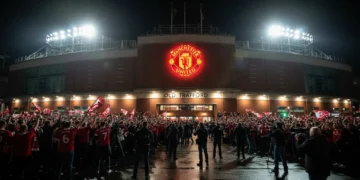 The Old Trafford stadium at night, with its red-brick facade and illuminated entrance, surrounded by excited fans and media. The Manchester United crest is prominently displayed, symbolizing the inten