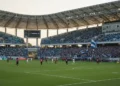 Gelora Bung Karno Stadium in Parepare is shown with excited fans in blue and white, cheering for PSM Makassar. The setting sun casts a golden glow over the playing field, highlighting the 1-0 lead on