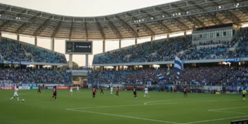 Gelora Bung Karno Stadium in Parepare is shown with excited fans in blue and white, cheering for PSM Makassar. The setting sun casts a golden glow over the playing field, highlighting the 1-0 lead on