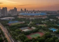 The scene shows the National Sports Complex in Jakarta, with a modern stadium illuminated by soft lighting. The stadium has distinctive arches and a bright blue roof, set against a backdrop of lush gr
