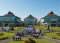 A vibrant and bustling campus of UMI Makassar, featuring the main building with its modern architecture and surrounded by green spaces. The scene showcases the 2,500 students participating in the Abdi